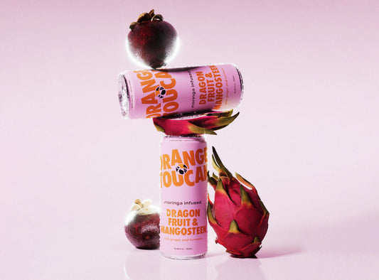 Two cans of Orange Toucan dragon fruit and mangosteen moringa infused drink, displayed with dragon fruit and mangosteen