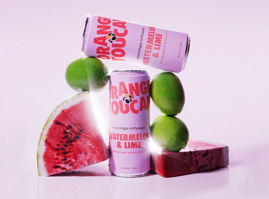 A can of moringa infused Orange Toucan watermelon and lime flavor, alongside slices of watermelon and whole limes against a light pink background.