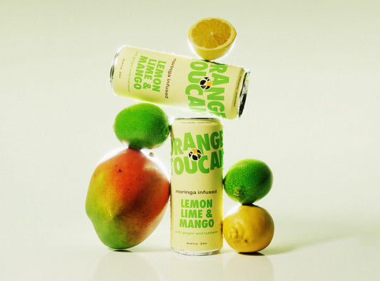 Two cans of lemon, lime, and mango flavored moringa infused Orange Toucan, pictured with a whole mango, one whole lemon, one half lemon and two limes.