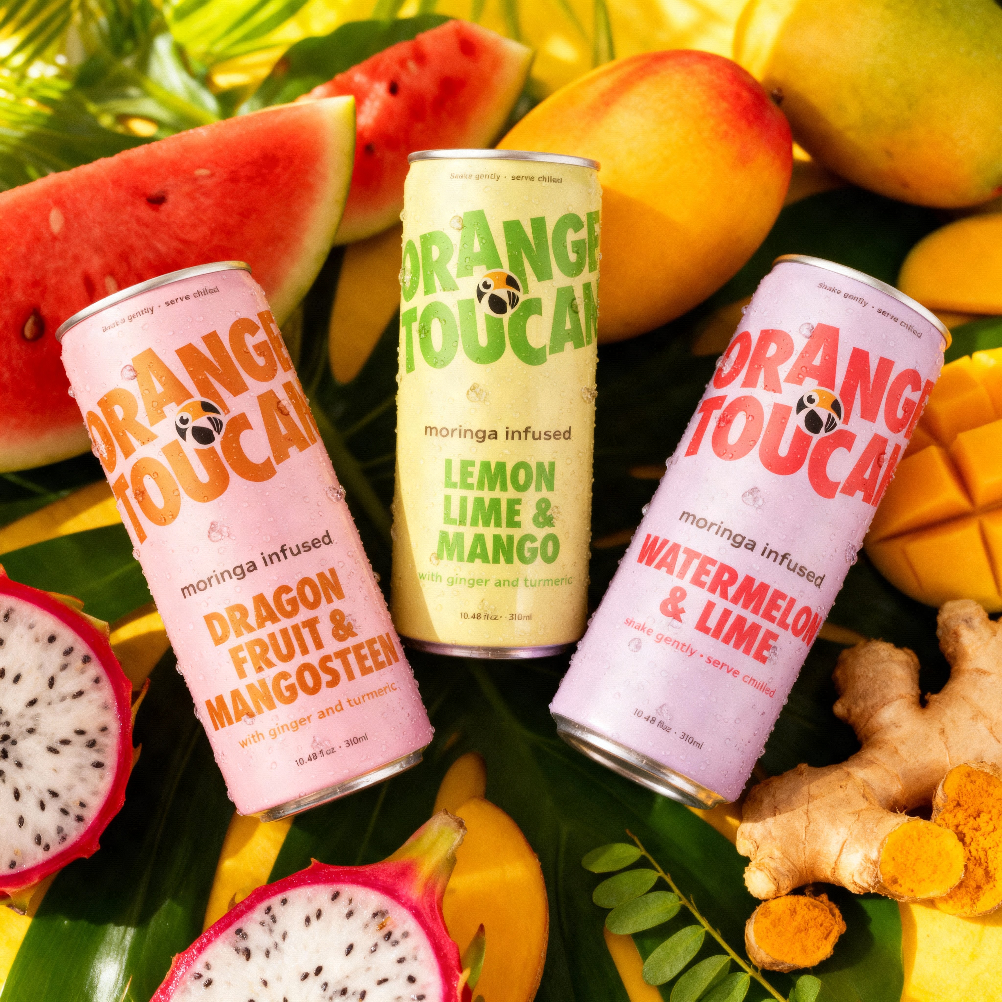 Three cans of Orange Toucan moringa infused drinks surrounded by mango, turmeric, watermelon, dragon fruit, and moringa leaves  on a leafy background