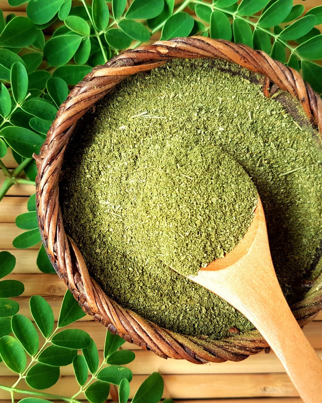 Green moringa powder in a woven basket with a wooden spoon, surrounded by green leaves on a bamboo mat.