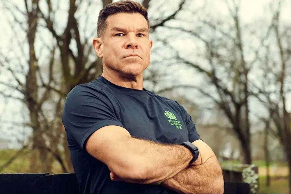 Ken Rideout wearing a dark blue t-shirt with a logo, standing outdoors with trees in the background